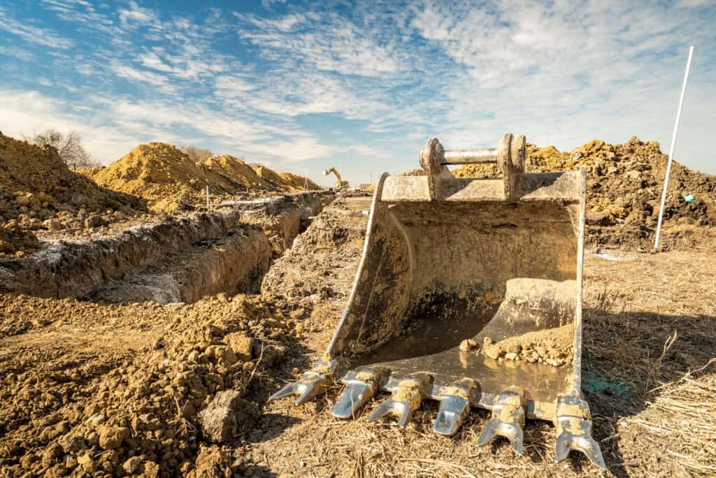 Excavator bucket digging into ground at trench site